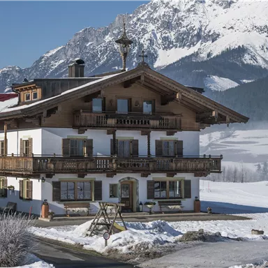 Ein traditionelles Alpenhaus im Schnee mit umliegenden Bergen. Die sonnige Landschaft vermittelt eine ruhige Winteratmosphäre.