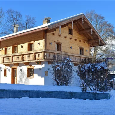 Ein traditionelles Holzhaus in einer schneebedeckten Landschaft. Im Hintergrund sind Bäume und Berge zu sehen.