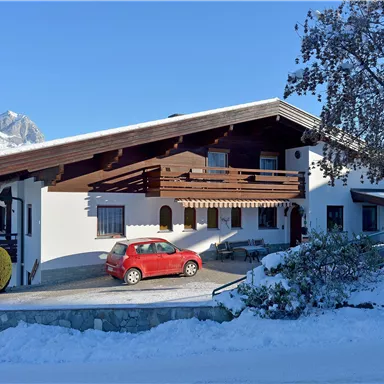 Ein schönes Haus im Schnee mit einem roten Auto davor. Im Hintergrund sind die Berge und ein klarer blauer Himmel sichtbar.