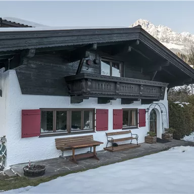 A charming alpine house with red shutters. Surrounded by snow-covered ground and mountains in the background.