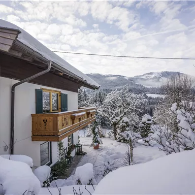 Ein charmantes Haus im Winter, umgeben von schneebedeckten Bäumen und Hügeln. Der Himmel ist klar mit einigen Wolken.