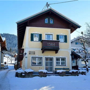 Ein charmantes, zweigeschossiges Haus im Schnee, umgeben von verschneiten Bäumen. Die Fassade ist traditionell mit Fenstern und einem Balkon gestaltet.