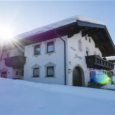 Ein charmantes, traditionelles Haus im Schnee, mit einem klaren blauen Himmel im Hintergrund. Die Sonne strahlt in das Bild und erzeugt eine warme Atmosphäre.