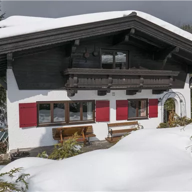 A charming Alpine house with red shutters, surrounded by snow. The winter magic is enhanced by the snowy landscape and the fir trees.