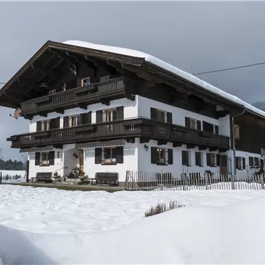 Ein traditionelles alpenländisches Haus mit mehreren Balkonen, umgeben von Schnee. Im Hintergrund sind Berge und ein bewölkter Himmel zu sehen.