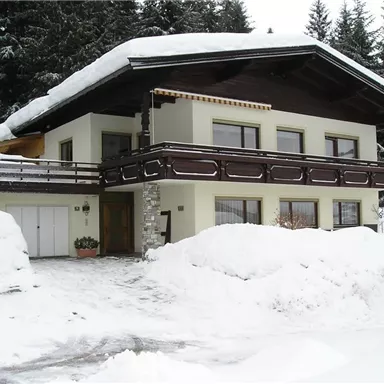 Ein charmantes Haus im Schnee mit einem großen Balkon. Umgeben von schneebedeckten Tannenbäumen.
