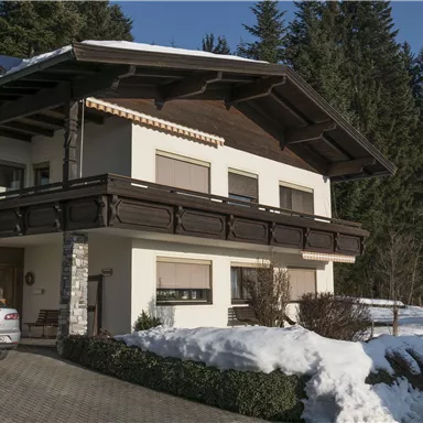 Ein modernes Haus im alpinen Stil mit einem Balkon und einer schneebedeckten Umgebung. Im Hintergrund sind grüne Bäume zu sehen.