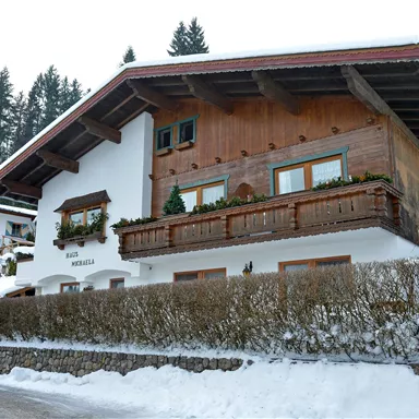 A cozy wooden house in the snow, surrounded by trees. The facade has large windows and a balcony.