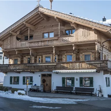 Ein traditionelles Holzhaus mit mehreren Balkonen und großen Fenstern. Es steht in einer schneebedeckten Landschaft und scheint in eine malerische Bergumgebung eingebettet zu sein.