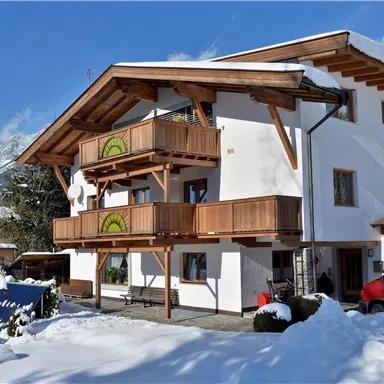 A beautiful old house with wooden balustrades, surrounded by snow. In the background, mountains and blue sky can be seen.