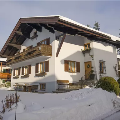 A traditional house in the snow with a steep roof and wooden balconies.  
Surrounded by a winter landscape and tall trees.