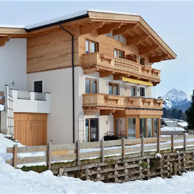 Ein modernes Chalet im Schnee mit Holzverkleidung und mehreren Balkonen. Im Hintergrund sind schneebedeckte Berge zu sehen.