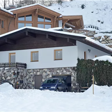 A house in the snow with a stone foundation and a modern roof. Fresh snow is everywhere, and there are some green bushes.