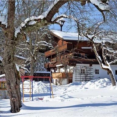 Ein schneebedecktes Haus umgeben von Bäumen. Im Vordergrund ist ein Spielplatz mit einer Schaukel zu sehen.
