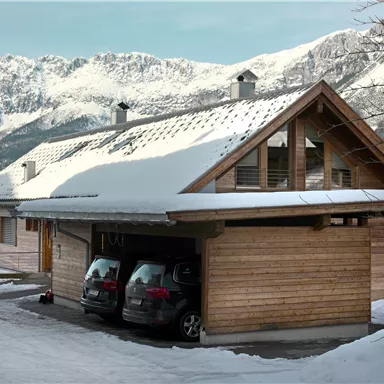 A modern wooden house in the snow, surrounded by mountains. Two cars are parked in the garage.