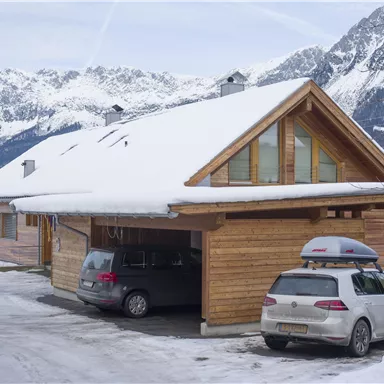 A cozy wooden house in the snow with a garage area. Impressive mountains can be seen in the background.
