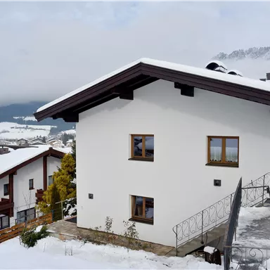 A modern house in the snow with a picturesque mountain landscape in the background. The windows offer a view of the snowy surroundings.