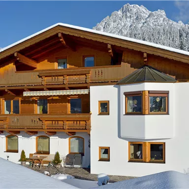 Ein chaletförmiges Haus aus Holz mit mehreren Balkonen steht im Schnee. Im Hintergrund sind schneebedeckte Berge zu sehen.