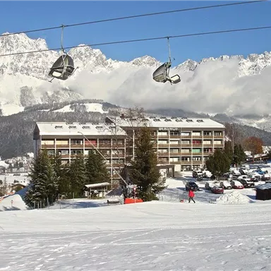 A snow-covered winter resort with a building and a cable car in the foreground. In the background, high mountains and a clear sky can be seen.