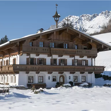 A traditional alpine house in the snow, surrounded by a winter landscape. The mountains are visible in the background and the sky is clear.