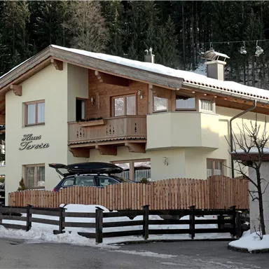 Ein gemütliches Haus im alpinen Stil mit Holzverkleidung und einem Balkon. Der Schnee und die umgebenden Bäume schaffen eine winterliche Atmosphäre.