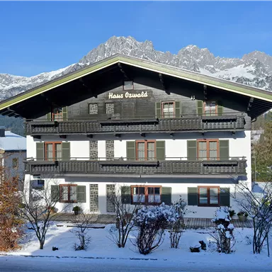 A traditional farmhouse surrounded by snow-capped mountains and trees. The sky is clear and blue, creating a peaceful atmosphere.