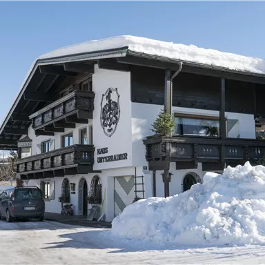 A traditional house in Alpine style, surrounded by snow. The facade is white with dark balconies and a stylish inscription.
