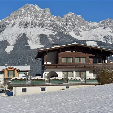 A picturesque winter landscape with snow-covered houses and impressive mountains in the background. The clear blue sky creates a tranquil atmosphere.