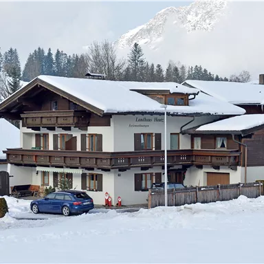 Ein gemütliches Haus im Schnee mit einem blauen Auto davor. Im Hintergrund sind schneebedeckte Berge und Nadelbäume zu sehen.