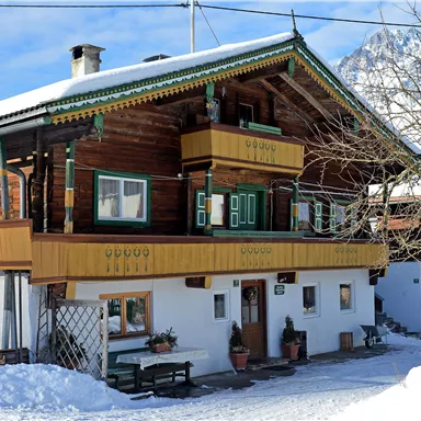 A traditional alpine wooden house in winter, surrounded by snow. Snow-covered mountains are visible in the background.