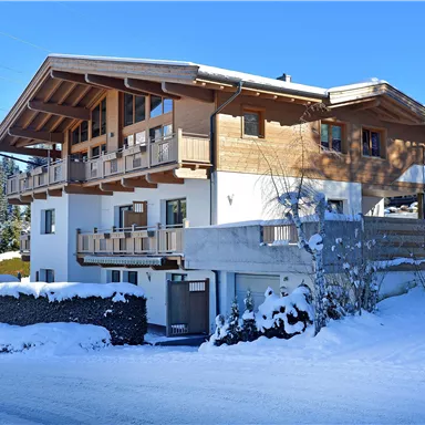Ein modernes, mehrstöckiges Holzhaus in einer verschneiten Landschaft. Umgeben von Bäumen und klarem blauen Himmel.
