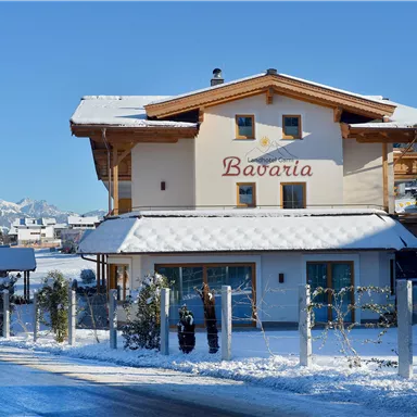 Ein gemütliches Holzhaus mit der Aufschrift "Bavaria" in einer verschneiten Landschaft. Im Hintergrund sind Berge und eine Kirche zu sehen.