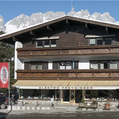 A traditional wooden building with a shop called Elektro Freydinger. In the background, there are snow-covered mountains and a clear blue sky.