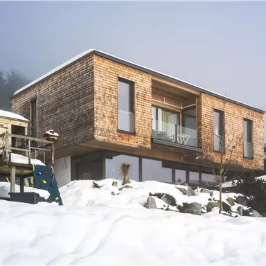 A modern wooden house surrounded by snow and fog. In the foreground, there is a playground with a seesaw.