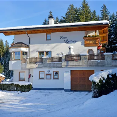A pretty white house in the snow, surrounded by fir trees. The balcony has a beautiful wooden cladding and there is a garage.