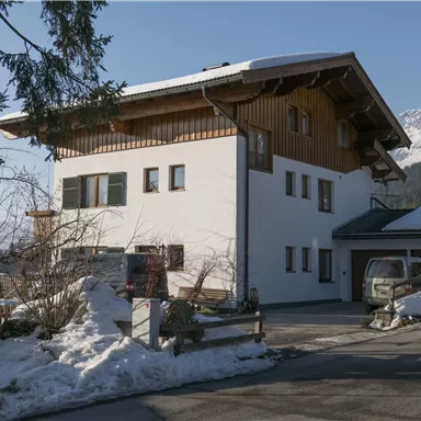 Ein modernes Haus mit Holzdetails, umgeben von schneebedecktem Boden und Bergen im Hintergrund. Der Himmel ist klar und die Umgebung wirkt ruhig.