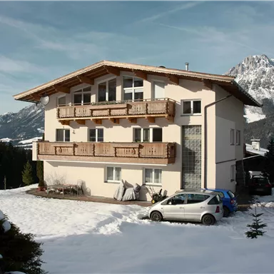 A large, modern house in the snow with a balcony and a view of the mountains. Several cars are parked around the house.