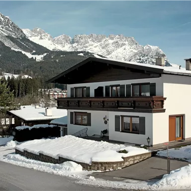 A modern house in the snow, surrounded by tall mountains. The sky is clear and the landscape seems calm and inviting.