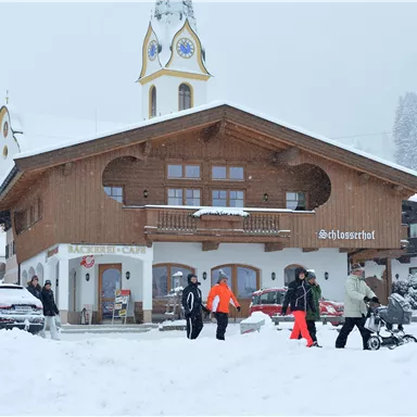 Ein traditionelles Holzhaus im Schnee mit einem Uhrenturm. Menschen gehen vorbei und genießen die winterliche Landschaft.