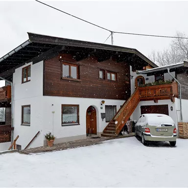 Ein gemütliches Holzhaus im Schnee, umgeben von einer winterlichen Landschaft. Es hat einen Eingang mit Treppe und ein Auto steht davor.