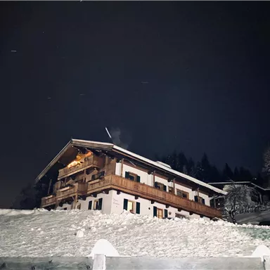 A cozy house in the snow at night. It is surrounded by trees and the landscape appears peaceful and wintry.