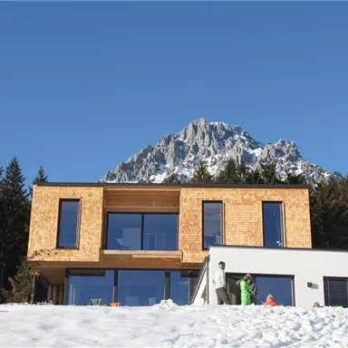 Ein modernes Haus aus Holz und Beton, umgeben von schneebedeckten Bergen. Der klare blaue Himmel verleiht der Szene eine ruhige Atmosphäre.