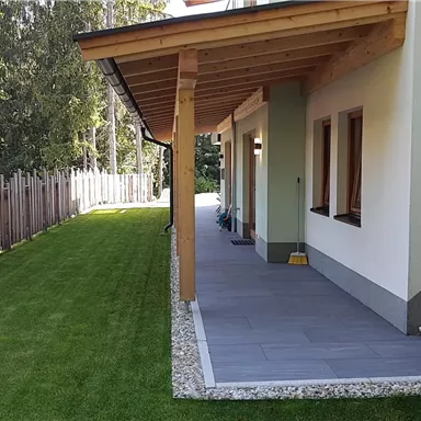 A modern house with a covered terrace and a well-kept garden. The path is designed with tiles and gravel and is surrounded by a slatted fence.