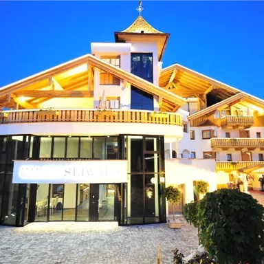 A modern hotel building with wooden cladding and a large glass entrance area. It is illuminated in the evening and is located in a quiet environment.
