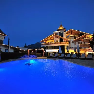 A luxurious hotel building with a illuminated facade and a large blue pool. The surroundings are elegant and welcoming.