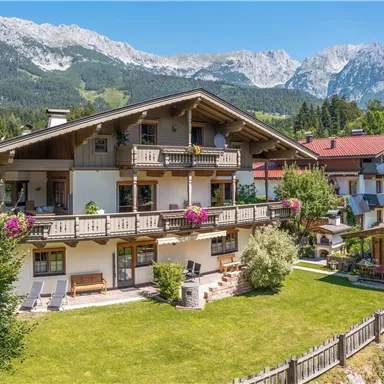 Ein malerisches Alpenhaus umgeben von grüner Wiese und Blumen. Im Hintergrund sind majestätische Berge zu sehen.