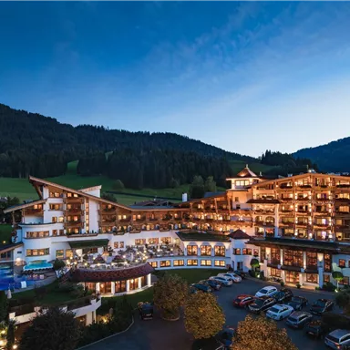 A beautiful hotel building with many lights at dusk. In the background, gentle hills and a clear sky can be seen.