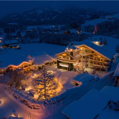 Ein malerisches Hotel in einer verschneiten Landschaft bei Nacht. Das Gebäude ist warm erleuchtet und von schneebedeckten Bäumen umgeben.