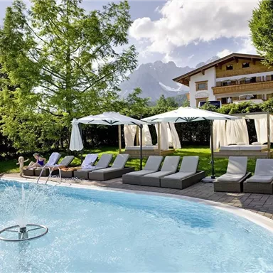 A beautiful pool area with sun loungers and cabanas in a green garden landscape. In the background, the mountains and a bright, blue sky can be seen.