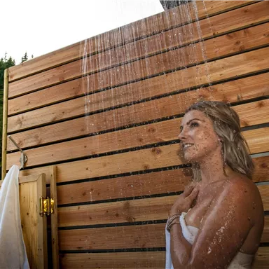 Eine Frau duscht unter freiem Himmel in einer Holzdusche. Im Hintergrund sind hohe Bäume zu sehen, die eine natürliche Umgebung schaffen.
